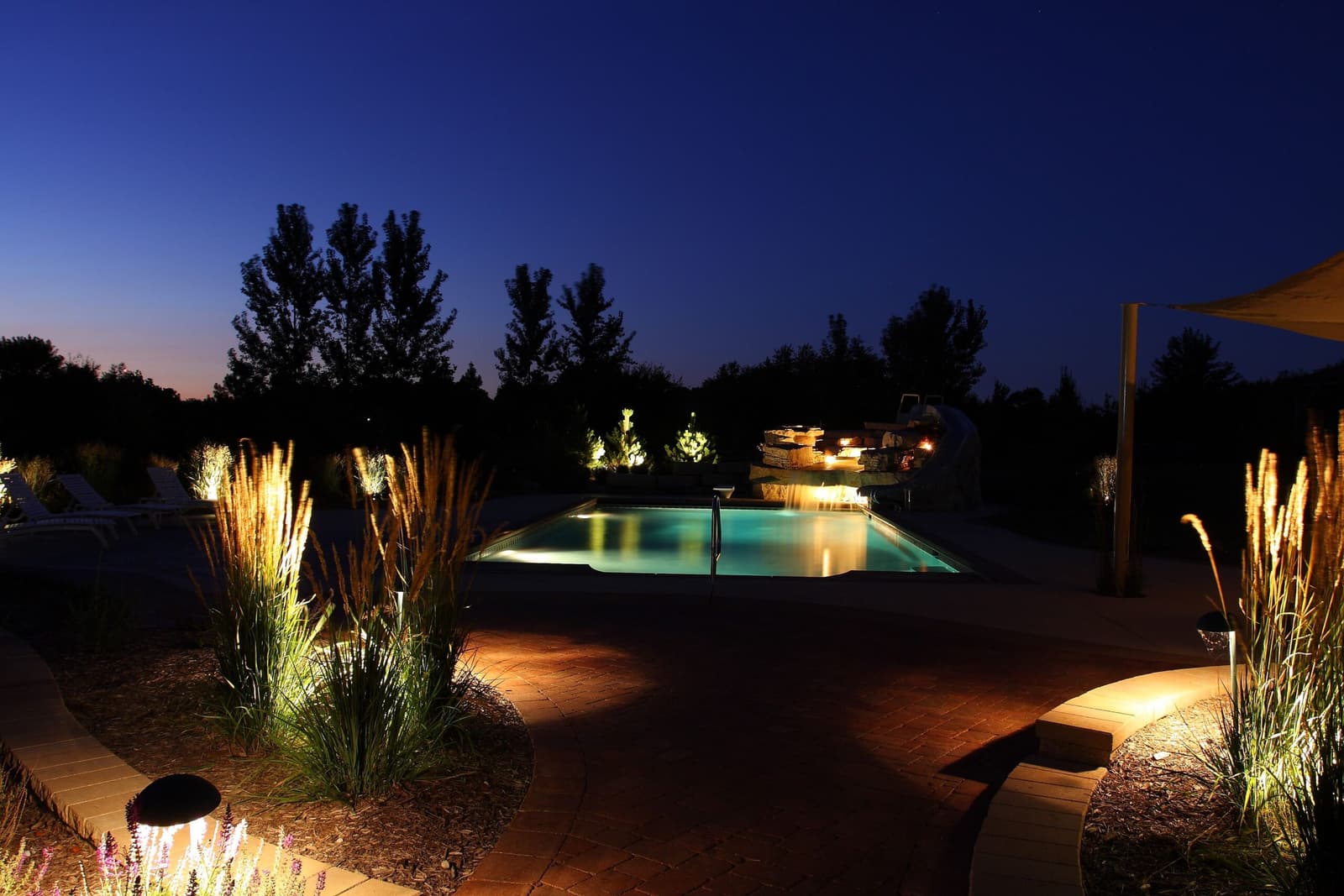 Pool glowing at dusk beyond landscape-lit ornamental grasses