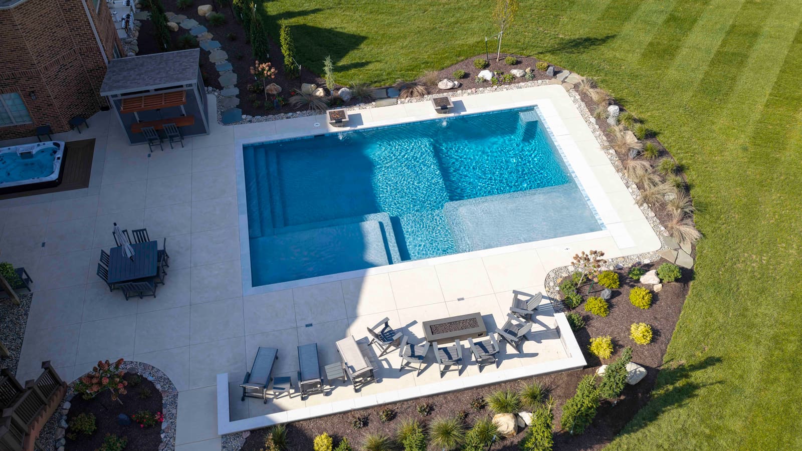 Overhead view of a rectangular gunite pool with a sun shelf, a wide white concrete deck, outdoor dining and lounge seating, and landscaped beds