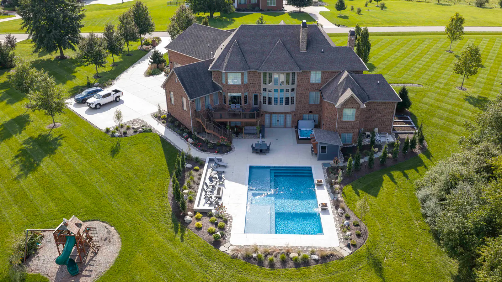 Wide aerial view of a brick home with a backyard gunite pool, curved landscape beds, and a play set on a striped lawn