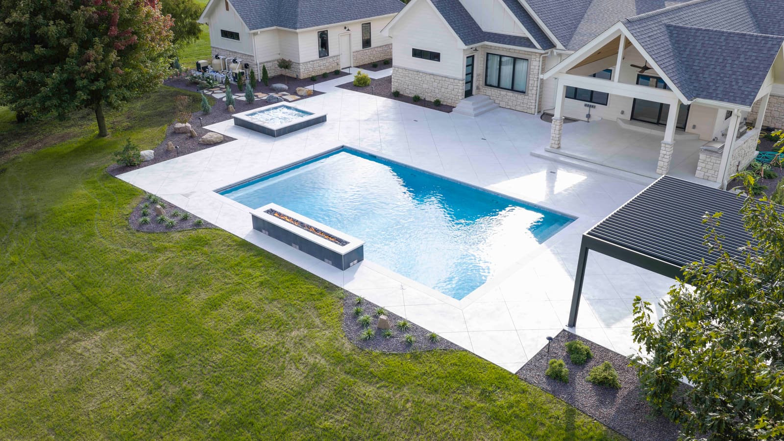 Aerial view of a rectangular gunite pool, raised square spa, and white paver patio beside a modern farmhouse