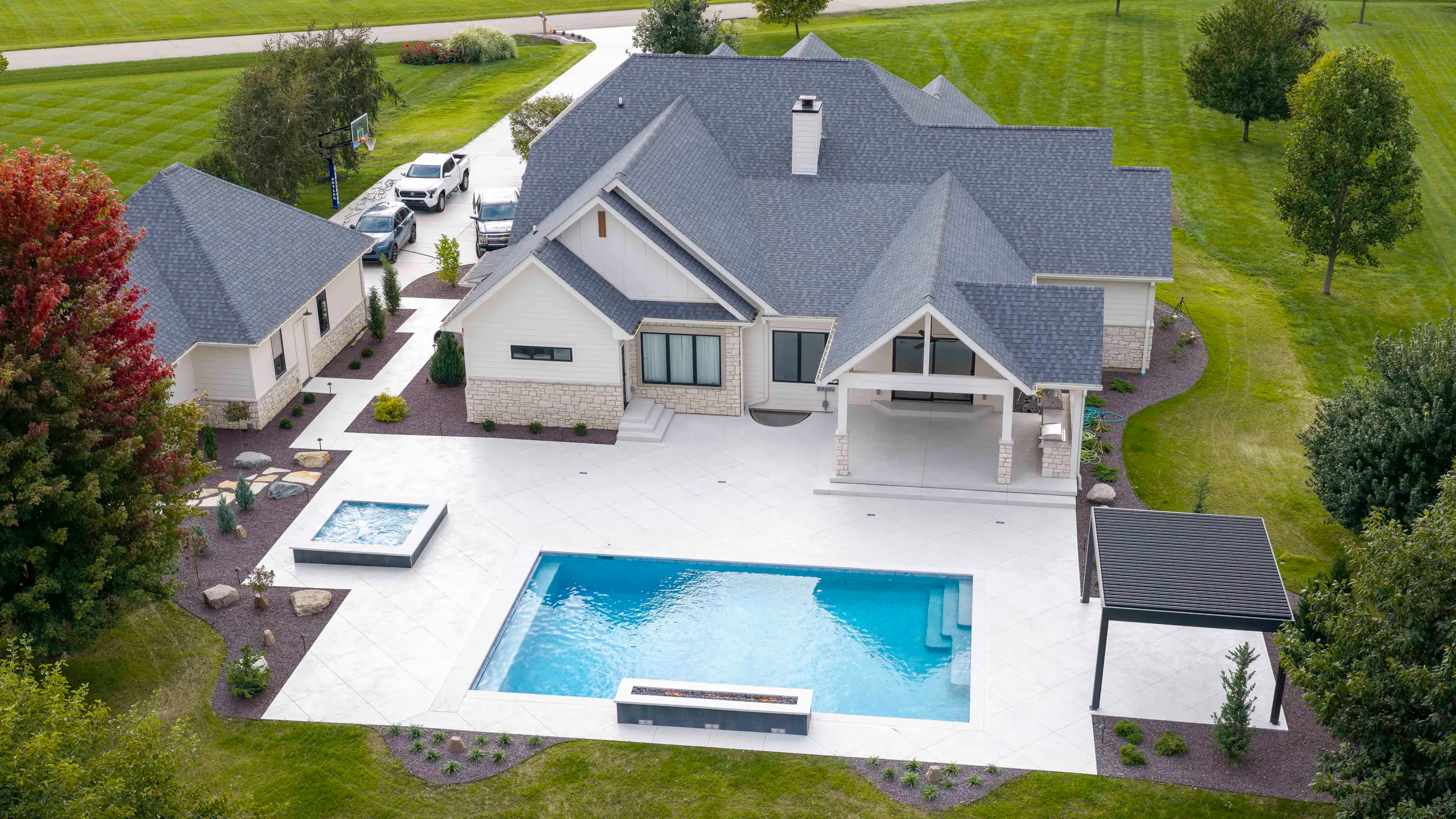 Aerial view of a modern farmhouse with a pool, raised spa, pergola, and white paver patio among mature trees