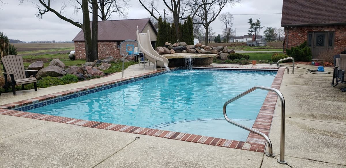 Pool with a brick-edged deck, handrail, and slide beside a brick home